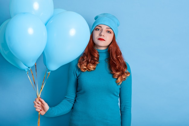 Traurige und gelangweilte junge europäische Frau, die Bündel von Heliumballons hält, während sie gegen blauen Raum stehen
