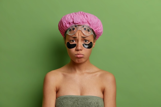 Traurige unglückliche junge afroamerikanische Frau hat schmollenden Gesichtsausdruck trägt Schönheitsflecken unter den Augen auf, um Schwellungen zu reduzieren, die in Badetuch gewickelt sind, das über grüner Wand isoliert wird
