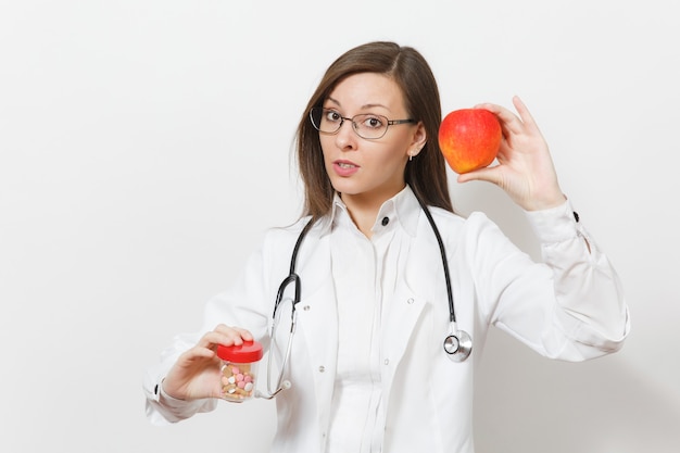 Traurige verärgerte hübsche junge Doktorfrau mit Stethoskop, Brille isoliert auf weißem Hintergrund. Ärztin im medizinischen Kittel hält Flasche mit Pillen, roter Apfel. Gesundheitspersonal, Medizinkonzept.