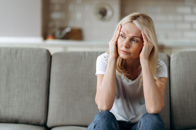 Foto traurige weiße ältere frau, die einfache kleidung trägt, sitzt allein zu hause auf der couch, fühlt sich unglücklich wegen kopfschmerzen, stress, krankheit oder schlechten nachrichten. sie braucht ruhe und schlaf.