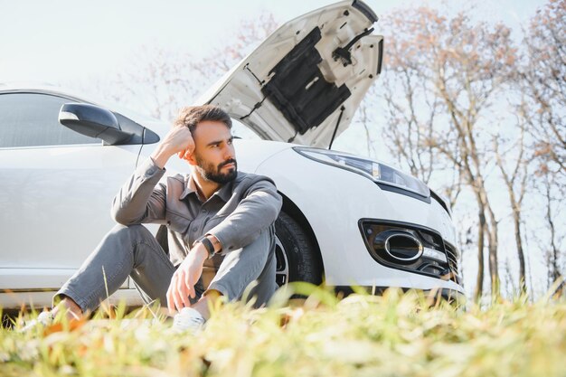 Trauriger Fahrer, der seinen Kopf hält und ein Motorproblem hat, das in der Nähe eines kaputten Autos auf der Straße steht Konzept der Autopanne