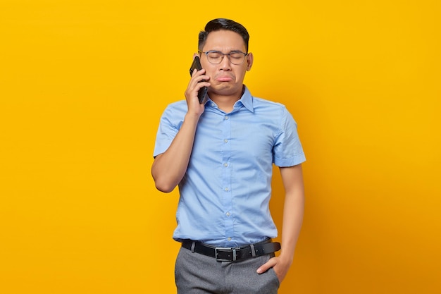 Trauriger junger asiatischer Mann Asiate mit Brille, der am Handy spricht, isoliert auf gelbem Hintergrund Geschäftsmann- und Unternehmerkonzept