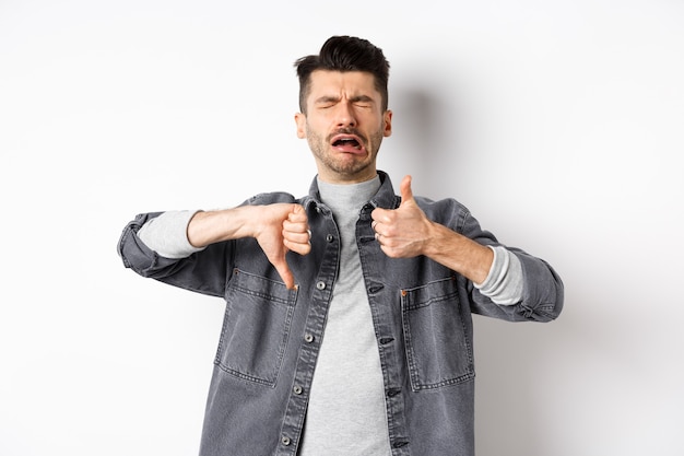 Trauriger Mann, der weint und Daumen nach oben zeigt, jammert und sich beschwert und enttäuscht vor weißem Hintergrund steht.