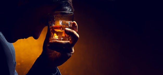 Trauriger Mann mit einem Glas Whisky im Gesicht. Whisky in einem Glas mit Eis, das Gesicht eines Mannes im Schatten