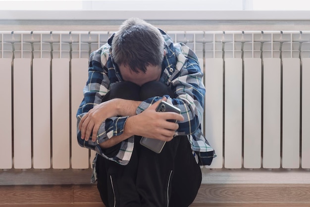 Trauriger Mann sitzt zu Hause auf dem Boden Das Konzept der Depression Die Heizung ausschalten Der Typ ist kalt und wärmt sich an der Batterie auf