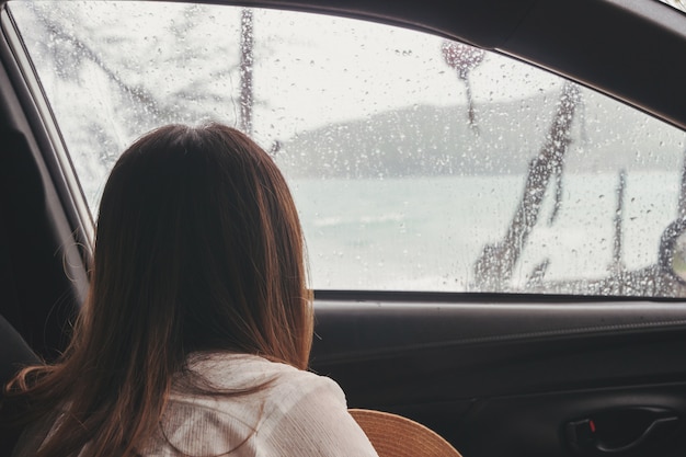 Trauriger reisender der jungen frau innerhalb des autos am regnerischen tag, der durch das fenster dem strand betrachtet