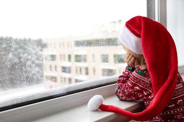 Trauriges, alleinstehendes Mädchen in weihnachtlicher roter Weihnachtsmütze blickt auf das Fenster und wartet auf die Feiertage Psychische Gesundheit