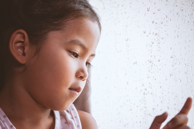 Trauriges asiatisches Kindermädchen, das draußen durch das Fenster am regnerischen Tag schaut