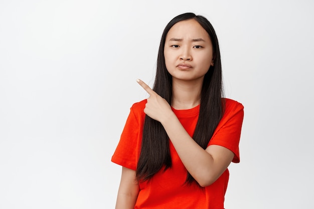 Trauriges asiatisches Mädchen, das mit dem Finger auf die obere linke Ecke zeigt und sich beschwert, schmollend launisch, in rotem T-Shirt auf Weiß stehend.