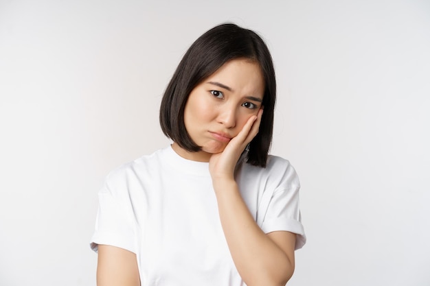 Trauriges asiatisches Mädchen, das verärgert und einsam aussieht, schmollend und stirnrunzelnd vor weißem Hintergrund in lässigem T-Shirt steht