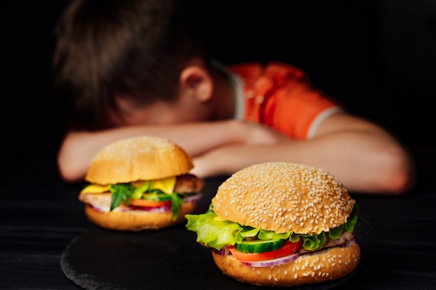 Trauriges Kind, das am Tisch sitzt und seinen Kopf auf Arme vor geschmackvollen Burgern setzt.