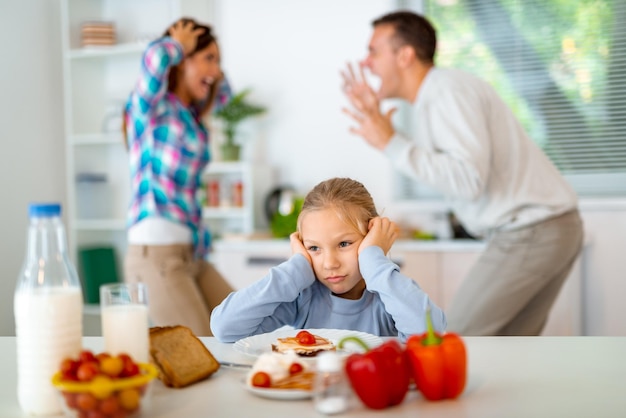 Trauriges kleines Mädchen beim Frühstück, während die Eltern in der Küche streiten. Sie ist verärgert, hält den Kopf und schaut weg.