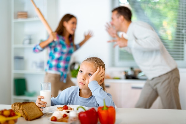 Trauriges kleines Mädchen beim Frühstück, während die Eltern in der Küche streiten. Sie ist verärgert, hält den Kopf und schaut weg.