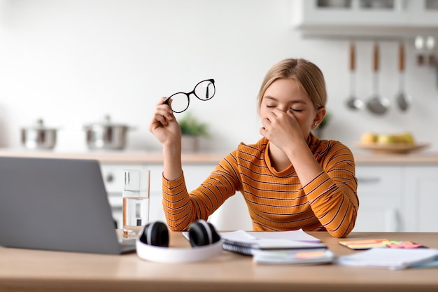 Trauriges müdes europäisches jugendlich blondes Mädchen nimmt Brille ab und reibt Augen am Tisch mit Laptop in der Küche