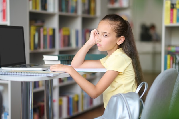 Trauriges müdes nachdenkliches süßes dunkelhaariges kaukasisches Schulmädchen, das am Tisch in einer öffentlichen Bibliothek sitzt