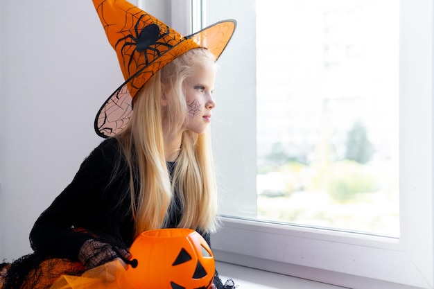 Trauriges, nachdenkliches blondes Mädchen im Hexenkostüm, das zu Hause im Fenster mit einem Süßigkeiteneimer schaut, der sich auf Halloween vorbereitet, um vor Coronavirus zu schützen
