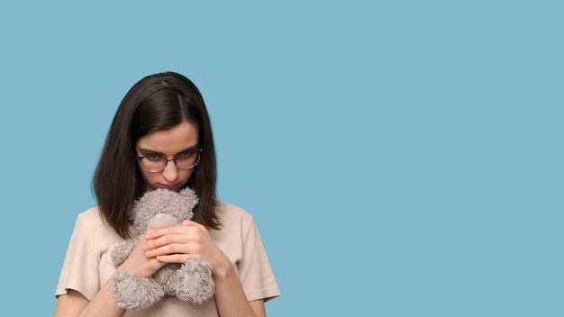 Trauriges, schönes Studentenmädchen mit Brille, das einen grauen Teddybären auf blauem Hintergrund umarmt