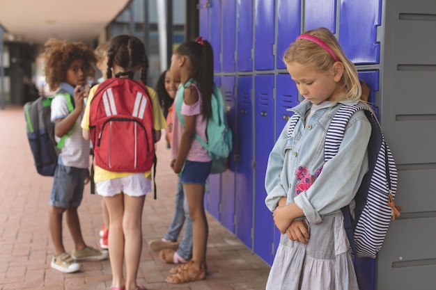 Trauriges Schulmädchen steht im Flur, während andere Schulkinder miteinander interagieren