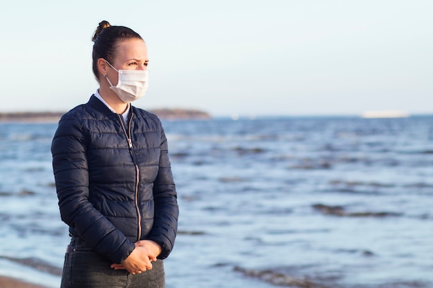 Trauriges unglückliches nachdenkliches frustriertes Mädchen, junge verärgerte verzweifelte Frau in der medizinischen Schutzmaske auf ihrem Gesicht gegen Coronavirus, das auf Strandmeer geht. Virus, Depression, Isolation, Epidemie, Drama-Konzept