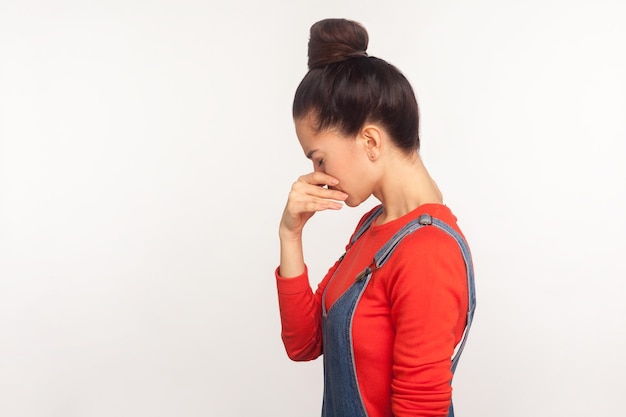 Traurigkeit und Depression Seitenansicht eines unglücklichen Mädchens mit Haarknoten in Denim-Overalls, das den Kopf beugt und weint, während es verzweifelte, traurige Gefühle erlebt, Studioaufnahme isoliert auf weißem Hintergrund