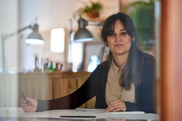 A través del cristal de una diseñadora haciendo bocetos en papeles mientras trabaja en el taller y mira la cámara