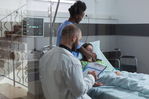 Foto a través de la ventana, la enfermera pone una máscara de oxígeno en una paciente inconsciente en una sala de hospital moderna. médico consultor escribiendo en el portapapeles con resultados de laboratorio médico mientras atiende el cuidador.