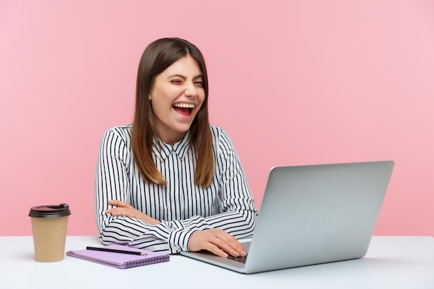 Traviesa y emocionada trabajadora de oficina con camisa a rayas sentada en el lugar de trabajo y guiñando un ojo mirando a la cámara con una sonrisa dentuda, buen humor, felicidad, tiro de estudio interior aislado en fondo rosa