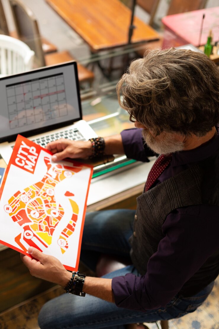 Foto trazando la ruta. hombre concentrado con un mapa de la ciudad en sus manos sentado con su computadora portátil en el café preparándose para su paseo.
