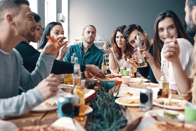 Treffen mit den besten Freunden. Gruppe junger Leute in Freizeitkleidung, die Pizza isst und lächelt, während sie eine Dinnerparty im Haus veranstalten