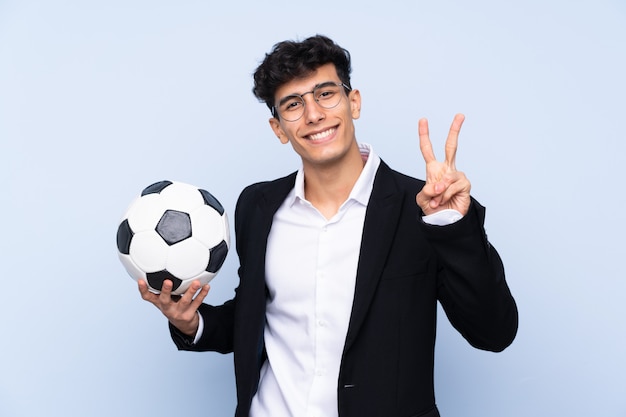 Treinador de futebol argentino sobre parede azul isolada, sorrindo e mostrando sinal de vitória