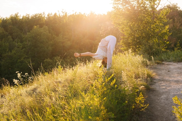 Treinador de ioga jovem em pé curva para frente com abridor de ombro à noite ao pôr do sol fêmea Yogini está realizando poses de ioga em cima do fundo rochoso da bela paisagem verde