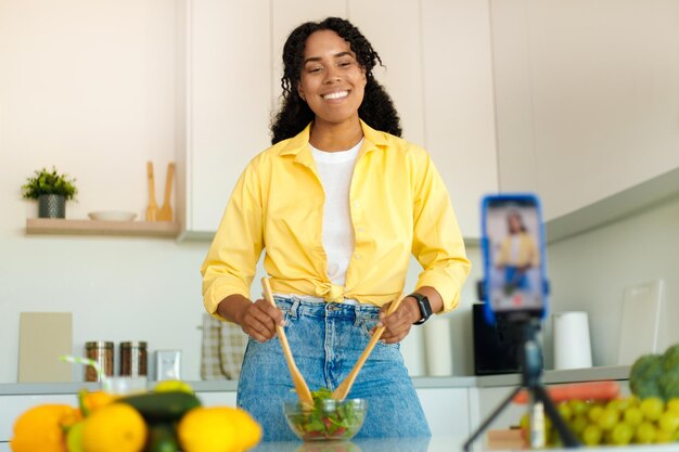 Treinador de nutrição Mulher negra animada gravando receita de vídeo na cozinha usando smartphone no tripé