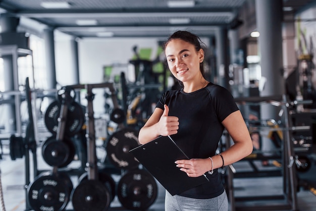 Treinador pessoal de fitness feminino jovem com bloco de notas em pé no ginásio com o polegar para cima.