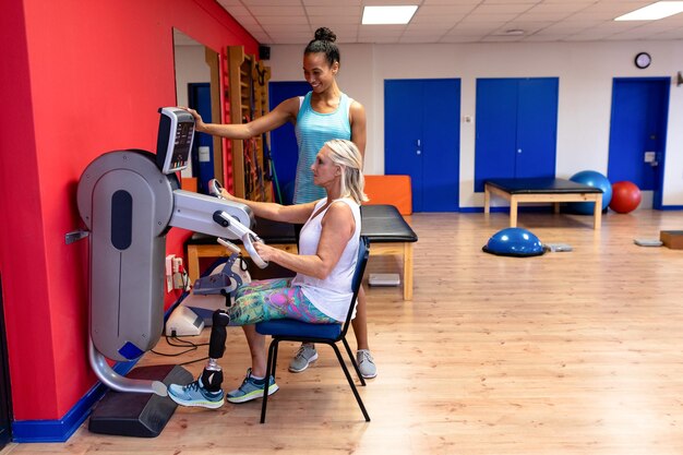 Foto treinadora assistindo uma mulher idosa com deficiência ativa a exercitar-se com equipamentos de exercícios