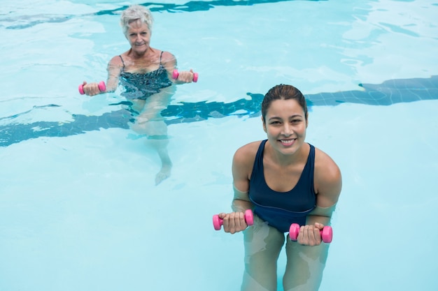 Treinadora e mulher sênior fazendo exercícios com halteres na piscina