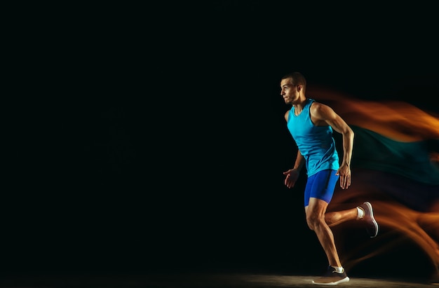 Treinamento de corredor masculino profissional isolado no fundo preto do estúdio em luz mista. Homem em traje esportivo, praticando corrida ou corrida. Estilo de vida saudável, esporte, treino, conceito de movimento e ação.