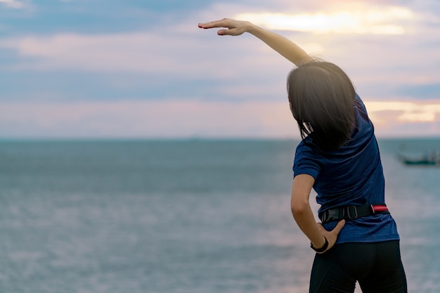 Treine a mulher da silhueta pela manhã na praia de pedra com o céu bonito do nascer do sol. Ajuste o corredor de mulher, esticando o corpo antes de executar. Cardio exercício para estilo de vida saudável.