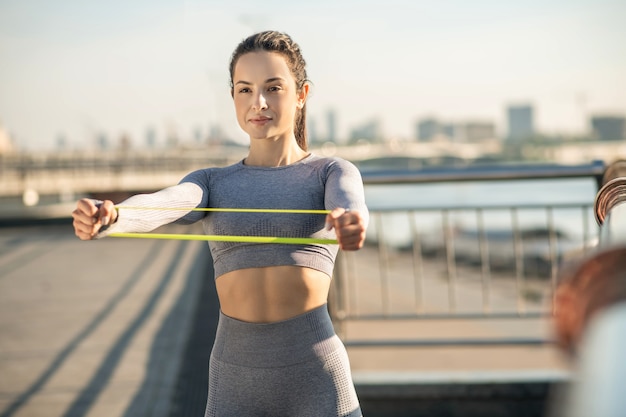 Treino de braços. Rapariga com roupa desportiva cinzenta a treinar os braços com uma faixa de resistência