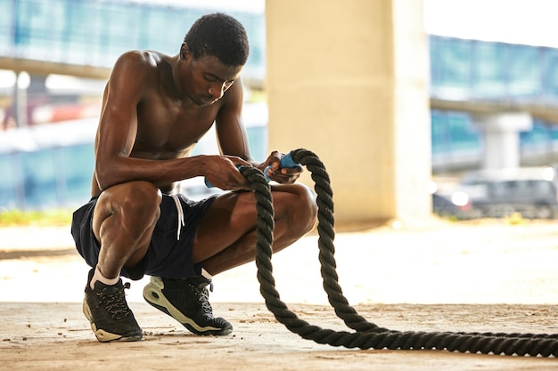 Treino de corda Homem esportivo fazendo exercícios de cordas de batalha ao ar livre Atleta masculino negro exercitando fazendo treinamento funcional com corda pesada