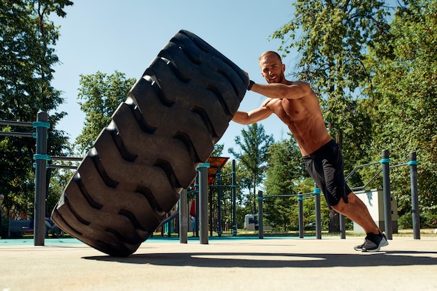 Treino de treinamento de homem forte, levantando pneu grande, ginásio ao ar livre, jovem adulto caucasiano esportivo, vira a roda grande na natureza, equipamento artesanal, esporte, exercitando o conceito de estilo de vida saudável