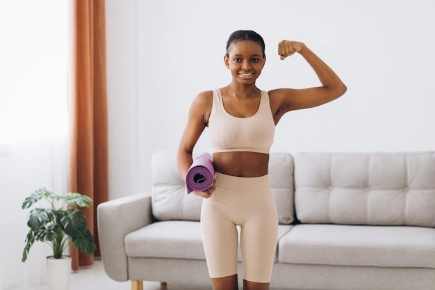 Treino doméstico Jovem negra desportiva segurando um tapete de ioga e mostrando o bíceps em casa treinando na sala de estar