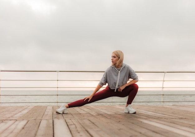 Treino matinal conceito de estilo de vida saudável mulher jovem e atraente em roupas esportivas faz alongamento das pernas antes do exercício na praia ao nascer do sol