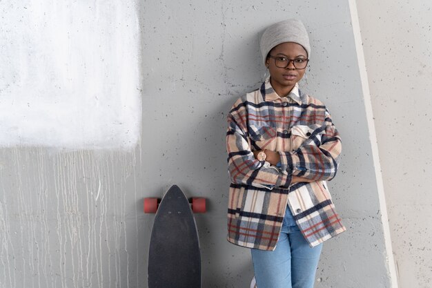 Trendige afrikanische Frau mit Skateboard junge schwarze Frau, die mit verschränkten Armen an der Betonwand steht