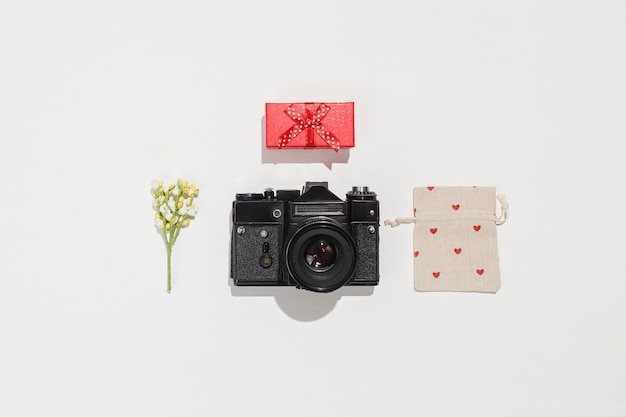 Trendige flache Laienkomposition mit Retro-Kamera, roter Geschenkbox, Geschenk-Leinentasche mit roten Herzformen und Frühlingsfeldblume auf weißem Hintergrund