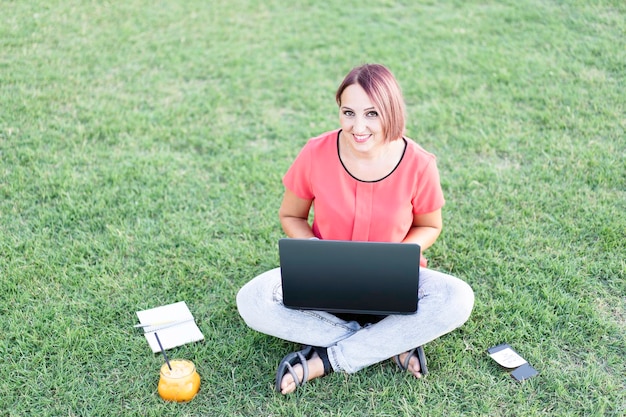 Trendige Frau, die im Freien mit Laptop arbeitet Lächelnde Frau, die auf dem Rasen sitzt und auf pc tippt Orangensaft ist bereit für die Pause Gesunder Lebensstil und Arbeiten im Freien Konzept mit Blick auf die Kamera copyspace