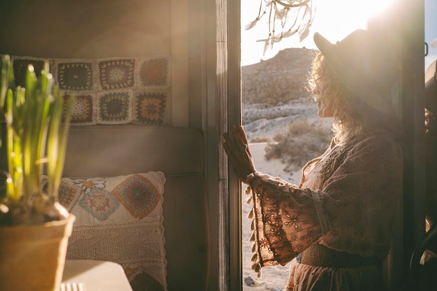 Trendige Frau im Boho-Kleid, die den Sonnenuntergang im Freien bewundert, der auf der Tür des Wohnmobils steht