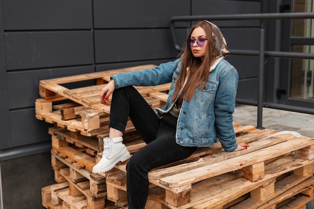 Trendige hübsche junge Hipsterin mit violetter Sonnenbrille in Militärkapuze in modischer Jeansjacke in Jeans in weißen Lederstiefeln entspannt sich auf Holzpaletten in der Stadt. Modernes Mädchenmodell im Freien.