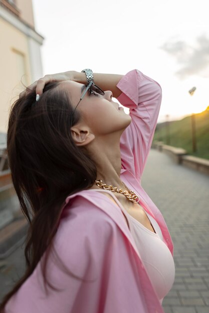 Trendige, schöne Modellfrau mit Vintage-Sonnenbrille und goldener Kette in rosafarbenem Hemd und Top-Spaziergängen und bei Sonnenuntergang im Freien genießen Frühlings-Sommer-Look-Kleidung im weiblichen Stil