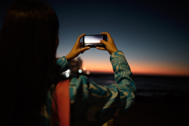 Trendige Touristin, die auf ihrem Handy ein Foto des Ozeans mit wunderschönem Sonnenuntergang am Horizont bei Nacht im Freien macht Influencer oder Vlogger, der ein Foto von der Natur mit Meerblick macht, um es in sozialen Medien zu posten oder zu teilen