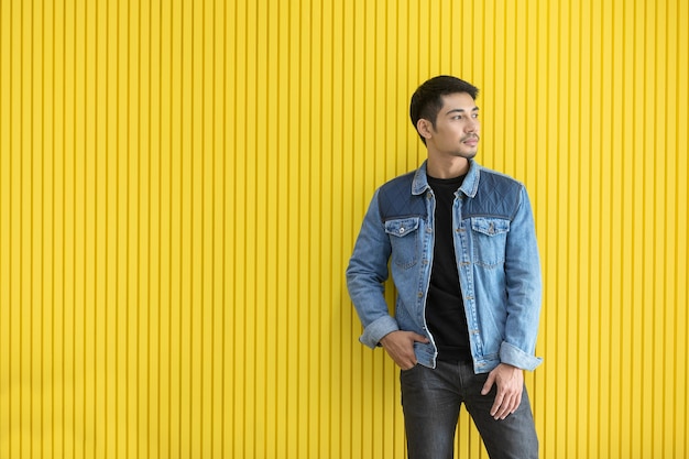 Trendiger attraktiver asiatischer Mann in Jeansjacke, die vor gelbem Hintergrund steht. Mittleres Porträt, modisches urbanes Konzept.
